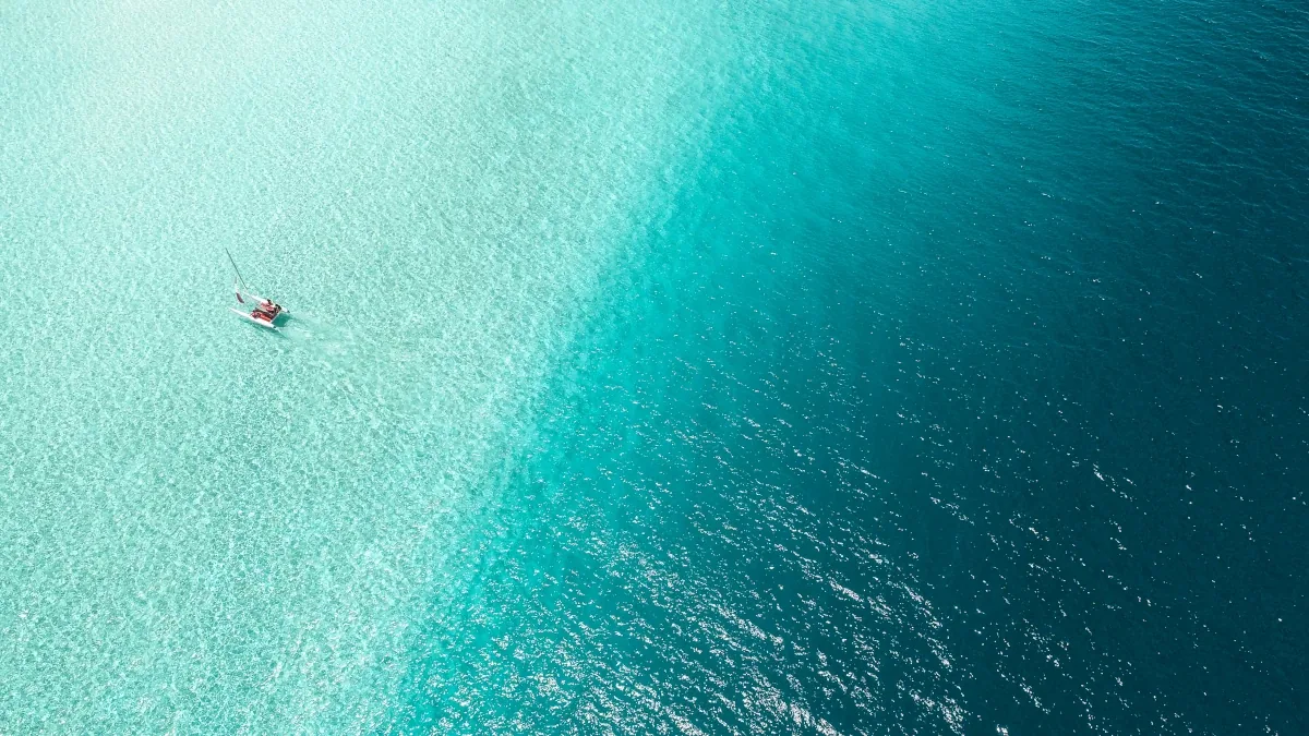 An aerial view of a small catamaran crossing the line where shallow turquoise water drops away into deep blue open sea.