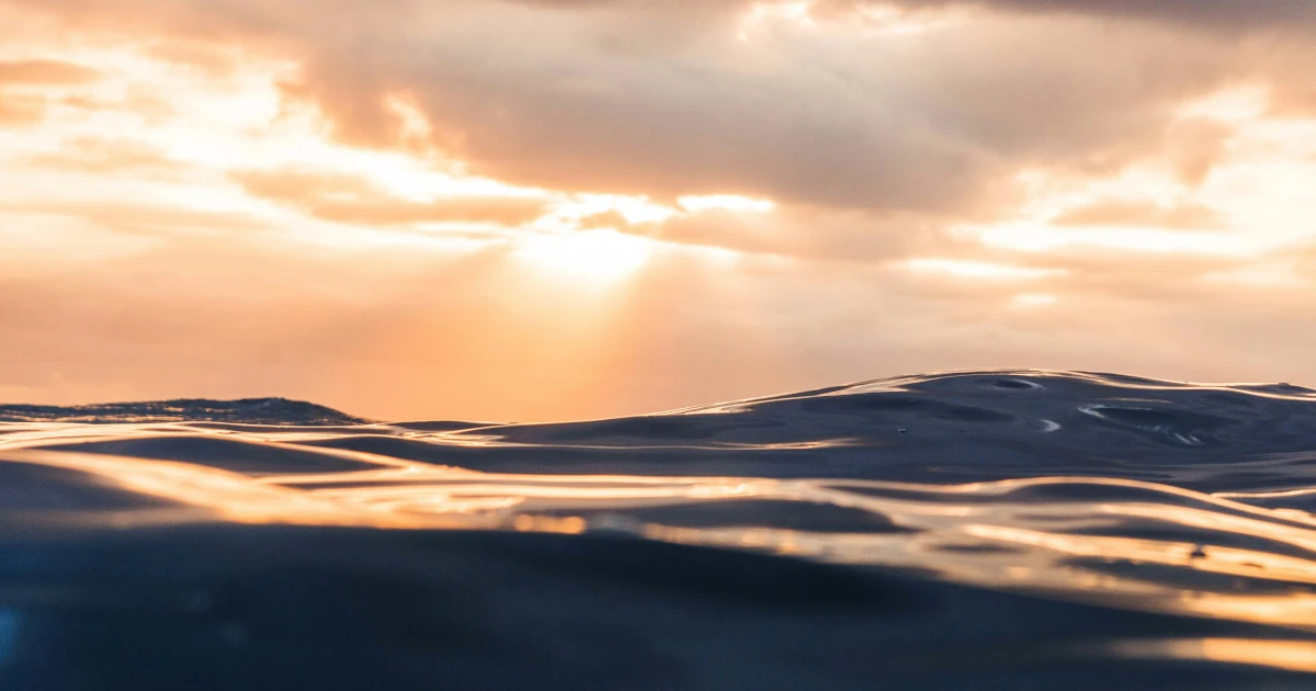 A low view across calm open water at sunset, the sun breaking through cloud above a gentle swell lit gold and blue.