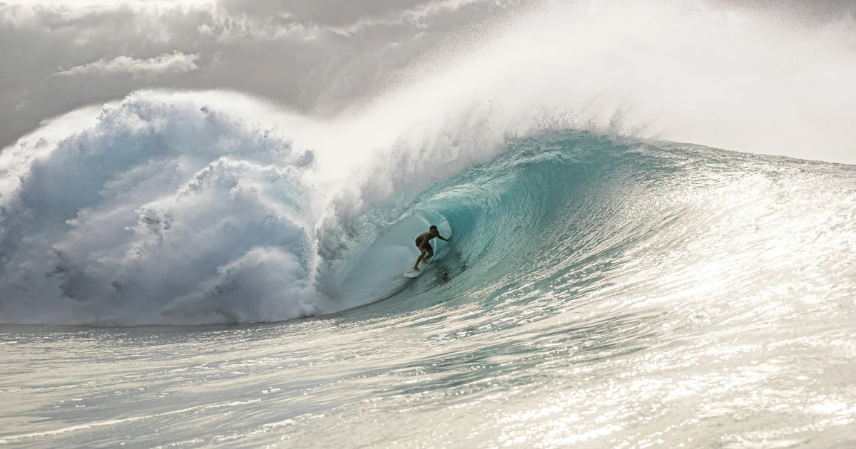 A surfer rides deep inside the barrel of a breaking turquoise wave under grey cloud, with spray blowing off the crest